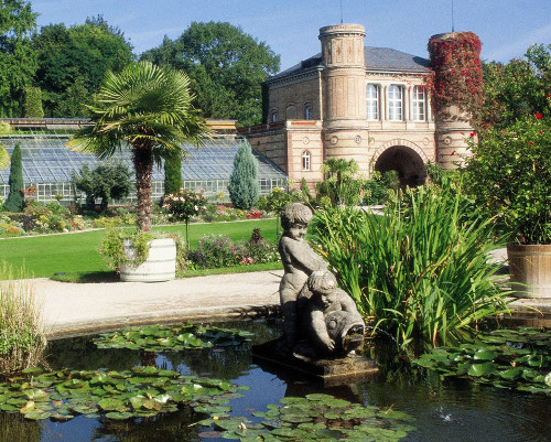 Eine Statue von zwei spielenden Kindern steht im Wasser eines Teichs mit Seerosen, umgeben von üppigen Pflanzen und einem historischen Gebäude im Hintergrund.